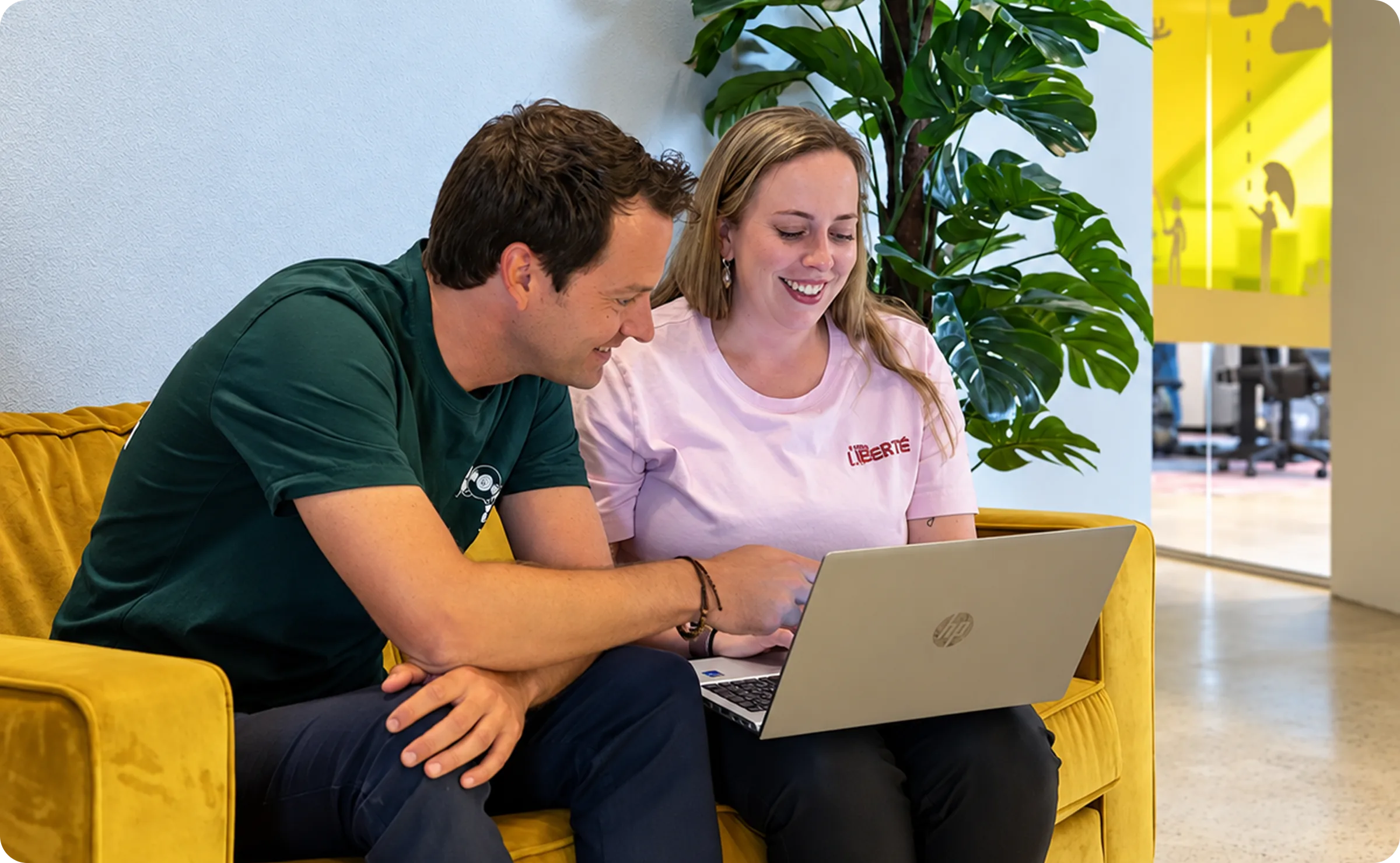 Twee mensen van DEREC kijken samen op een laptop, zittend op een gele bank
